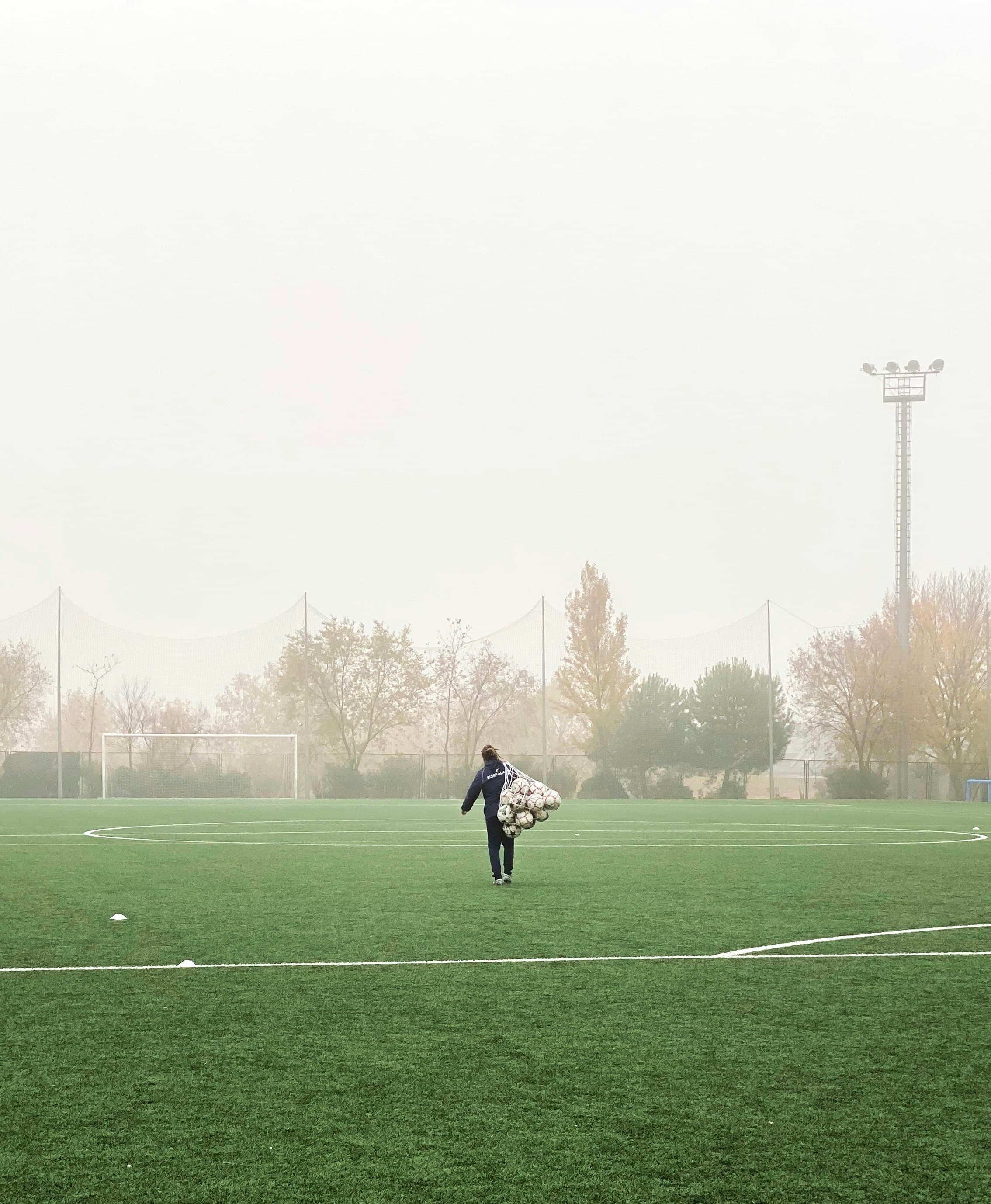 Coach carrying a bag of soccer balls across a foggy pitch